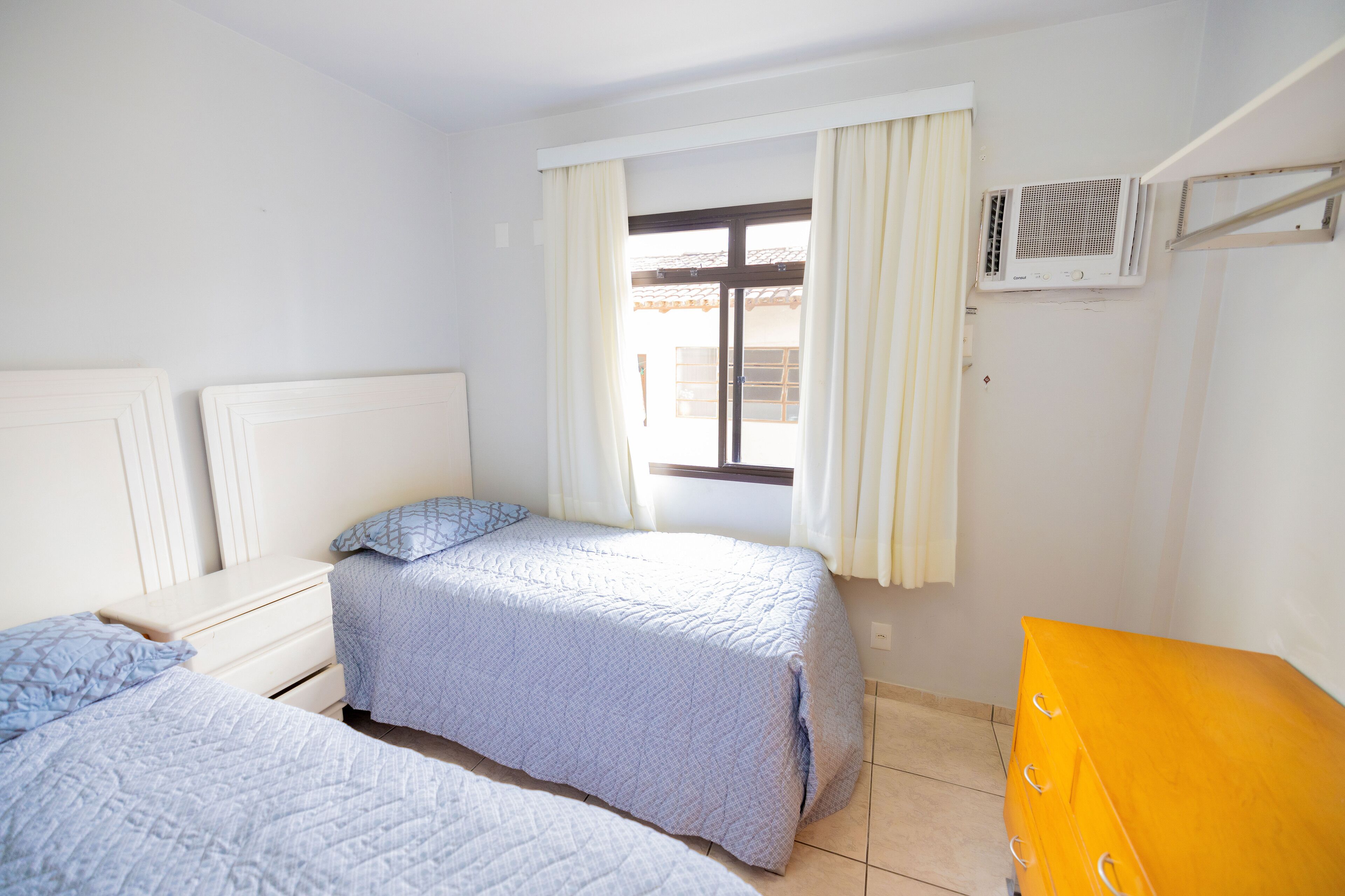 A bedroom with two beds, a window with curtains, an air conditioning unit, and a wooden dresser.