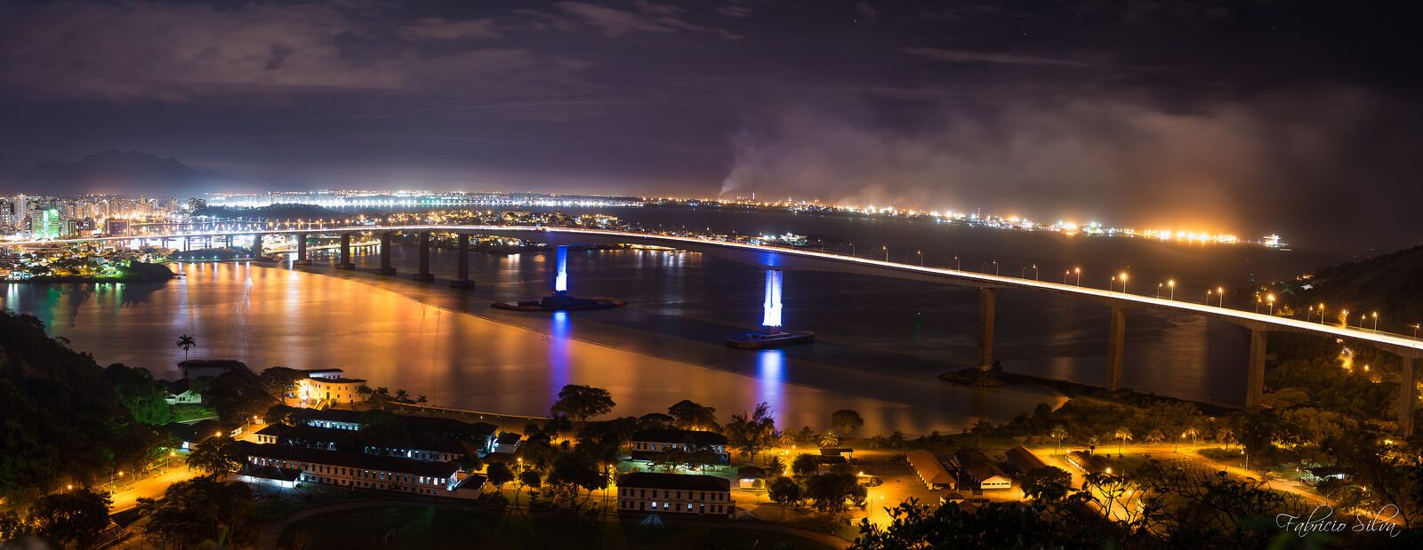 Vista nocturna de un paisaje urbano con un puente destacado, fuentes iluminadas y un horizonte lejano.
