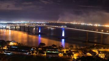 Vista nocturna de un paisaje urbano con un puente destacado, fuentes iluminadas y un horizonte lejano.