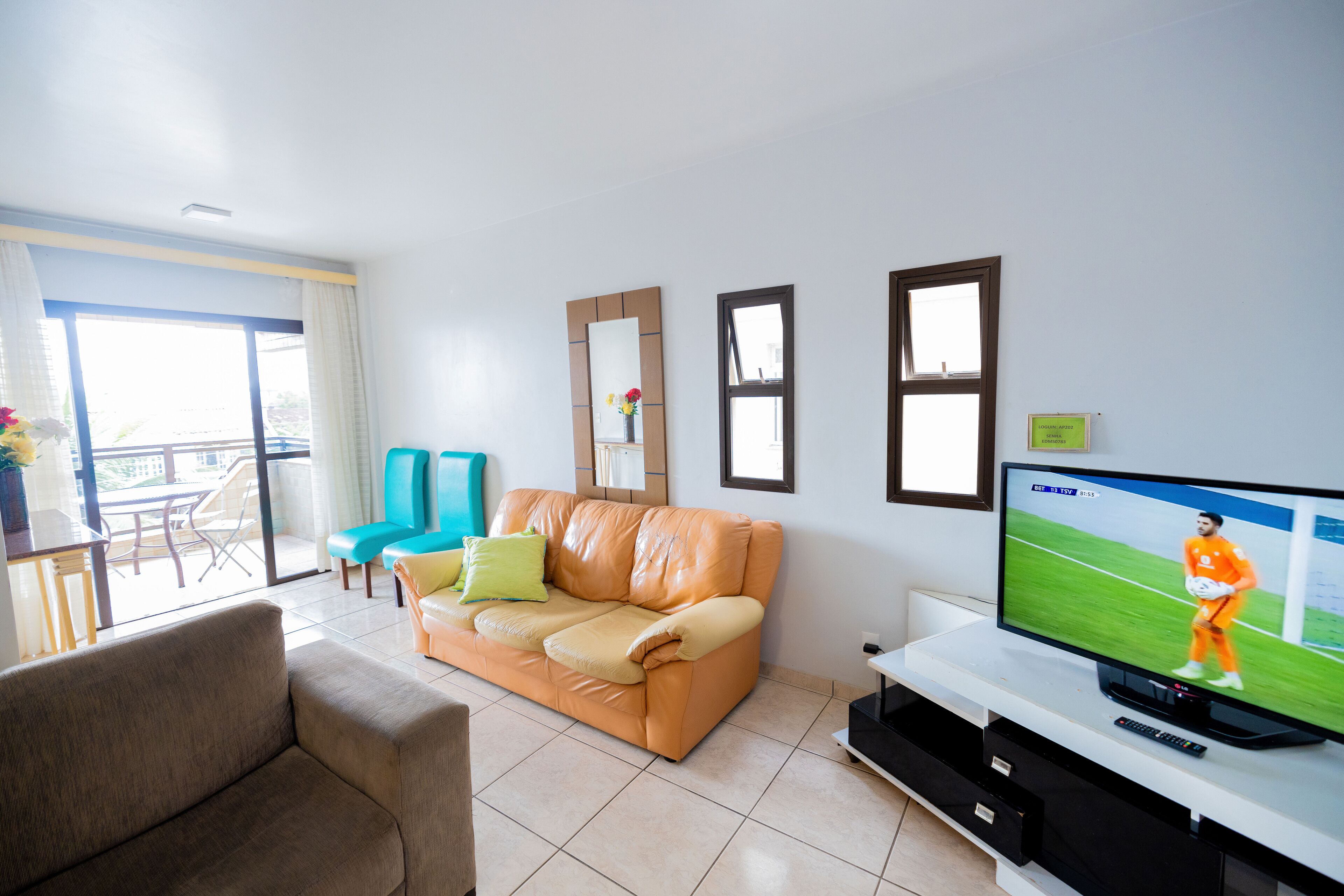 A living room with a sofa, a television displaying a sports event, and a balcony with a table and chairs.