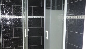 A glass-enclosed shower with a white frame, black tiled walls, and a small trash bin to the left. - Phoenix Hôtel (Béziers)