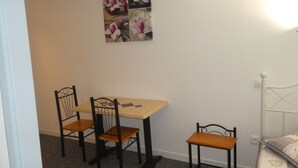 A small room with a wooden table and two chairs, a bed with a patterned blanket, and a wall-mounted picture of flowers. - Phoenix Hôtel (Béziers)