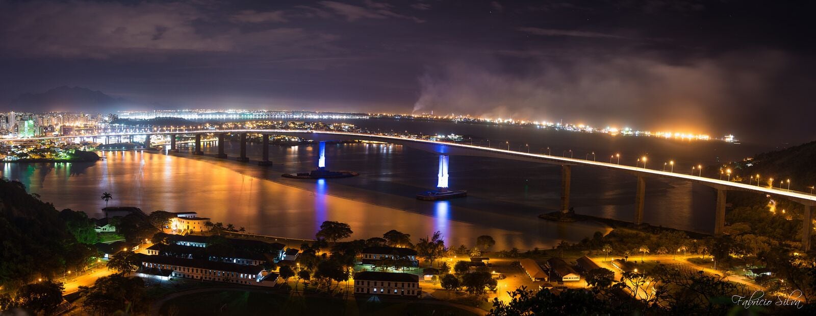 A nighttime view of a cityscape with a prominent bridge, illuminated water features, and a distant city skyline.