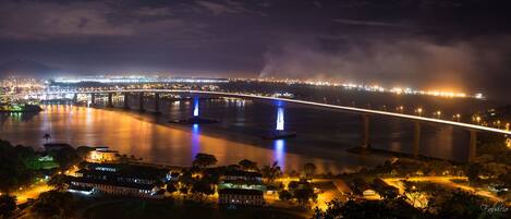 A nighttime view of a cityscape with a prominent bridge, illuminated water features, and a distant city skyline.