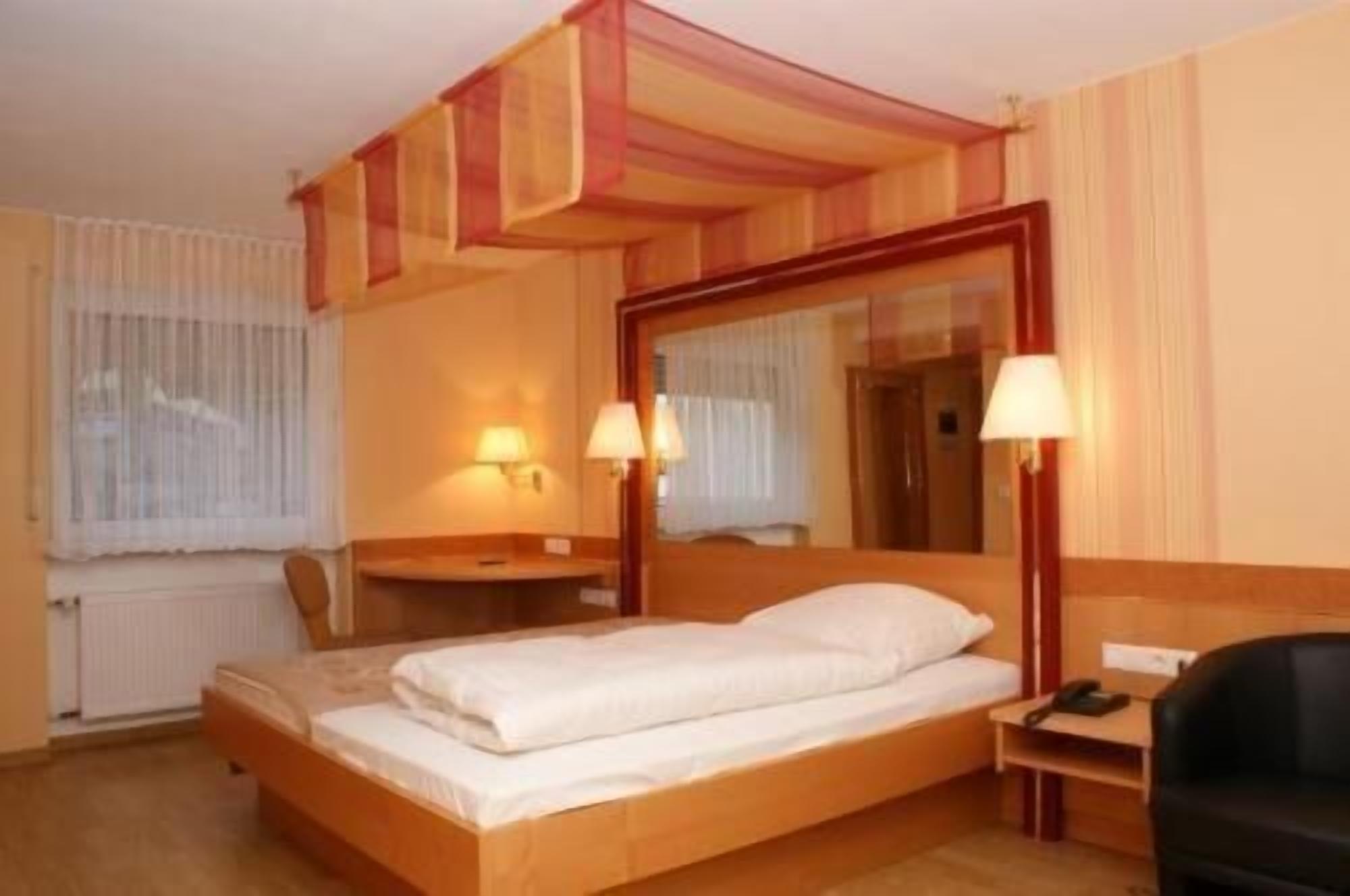 A hotel room with a four-poster bed, two bedside lamps, a small bedside table, and a chair.