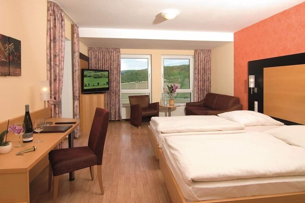 A hotel room with a bed, a desk with a chair, a TV, and a window with a view of greenery.