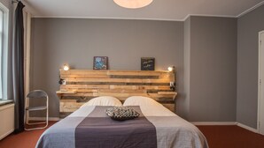 Une chambre à coucher avec une tête de lit en bois, un lit recouvert d’une couverture grise et blanche, deux oreillers, une petite étagère avec des livres et une chaise.