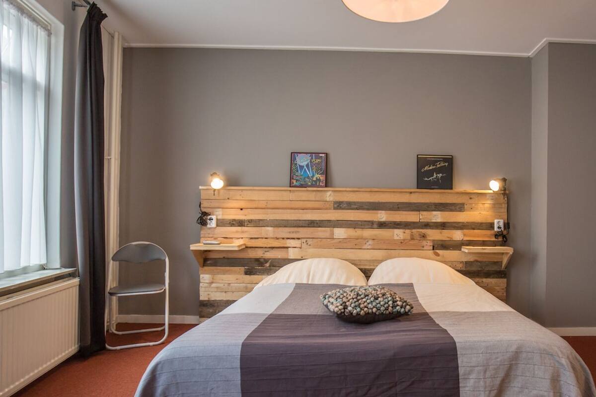 A bedroom with a wooden headboard, a bed with a gray and white bedspread, two pillows, a small shelf with books, and a chair.