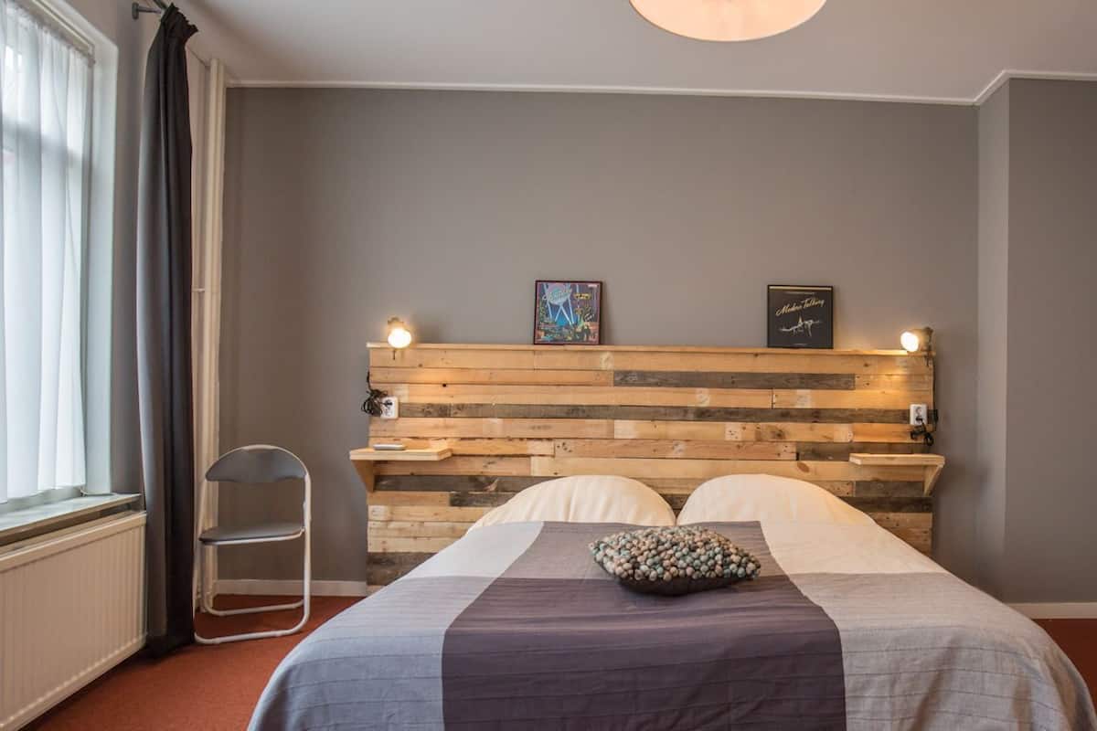 A bedroom with a wooden headboard, a bed with a grey and white bedspread, two pillows, a small shelf with framed pictures, and a window with curtains.