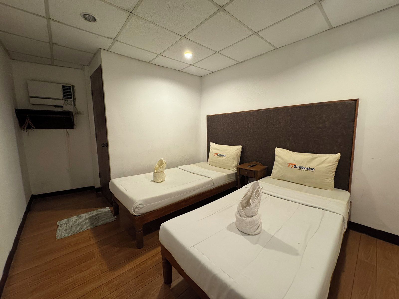 A hotel room with two beds, each with a pillow and a towel. There is a wooden floor, a ceiling light, and an air conditioning unit on the wall.