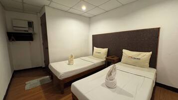 A hotel room with two beds, each with a pillow and a towel. There is a wooden floor, a ceiling light, and an air conditioning unit on the wall.