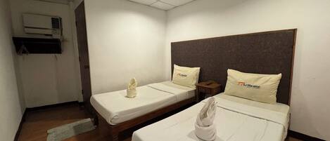 A hotel room with two beds, each with a pillow and a towel. There is a wooden floor, a ceiling light, and an air conditioning unit on the wall.