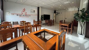A spacious cafe interior with multiple wooden tables and chairs, a large plant, and a wall-mounted sign reading "Hugs & Beans Specialty Coffee". - Sj Mansion Hotel (San Jose)
