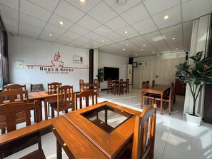 A spacious cafe interior with multiple wooden tables and chairs, a large plant, and a wall-mounted sign reading "Hugs & Beans Specialty Coffee". - Sj Mansion Hotel (San Jose)