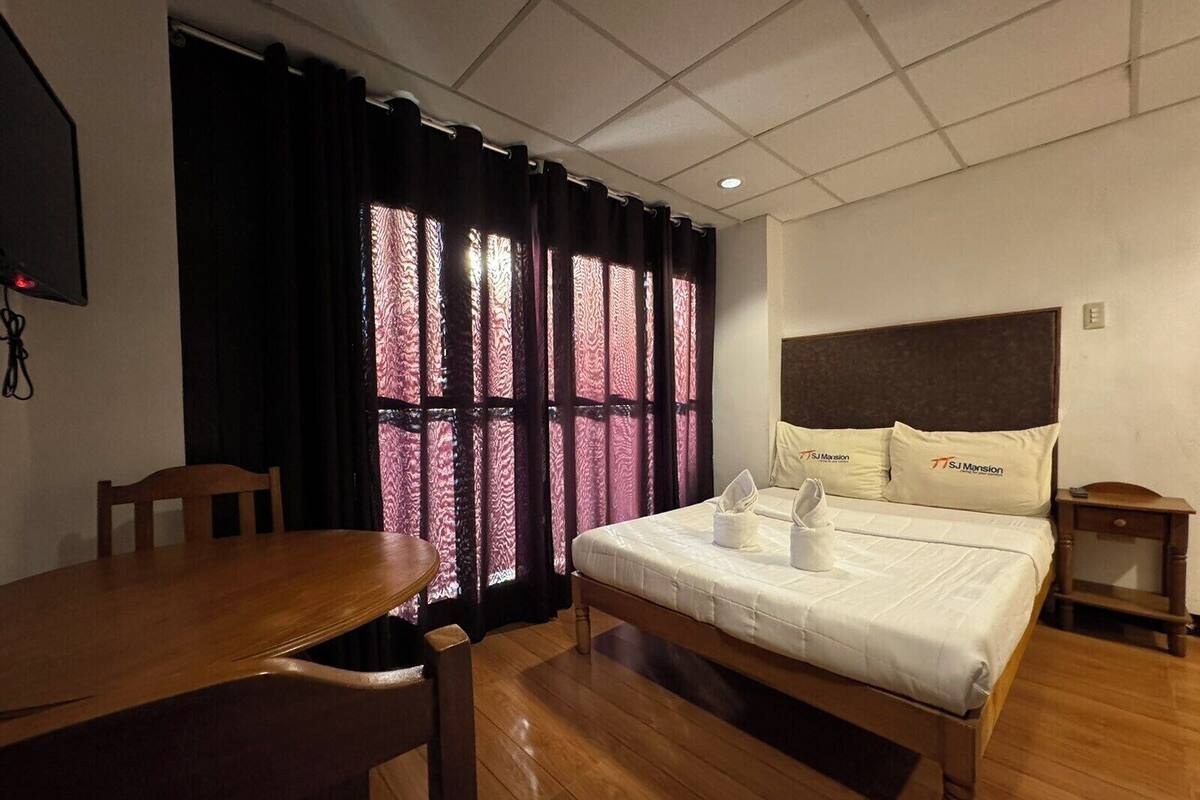 A hotel room with a wooden bed, a wooden table, and a window with patterned curtains.