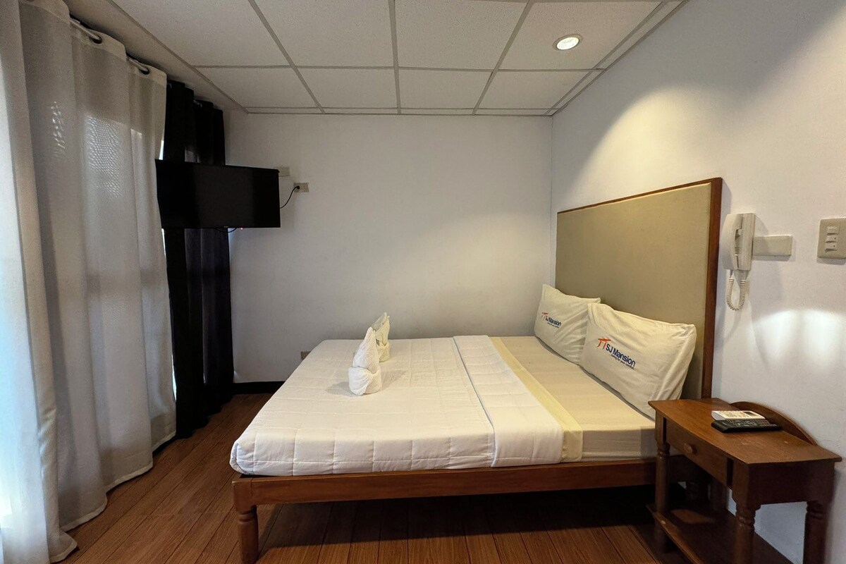 A hotel room with a wooden bed, white bedding, a bedside table with a phone, and a TV mounted on the wall.