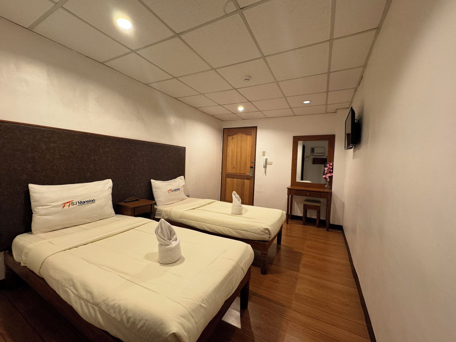 A hotel room with two beds, a wooden headboard, a small desk, a mirror, and a TV on the wall.