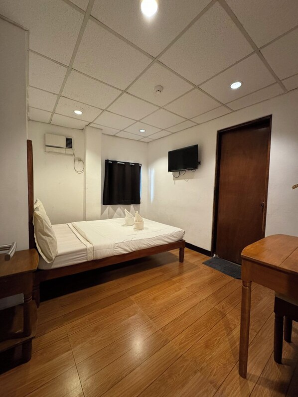 Quarto de hotel com uma cama de solteiro, uma mesa de madeira, televisão na parede e uma porta.