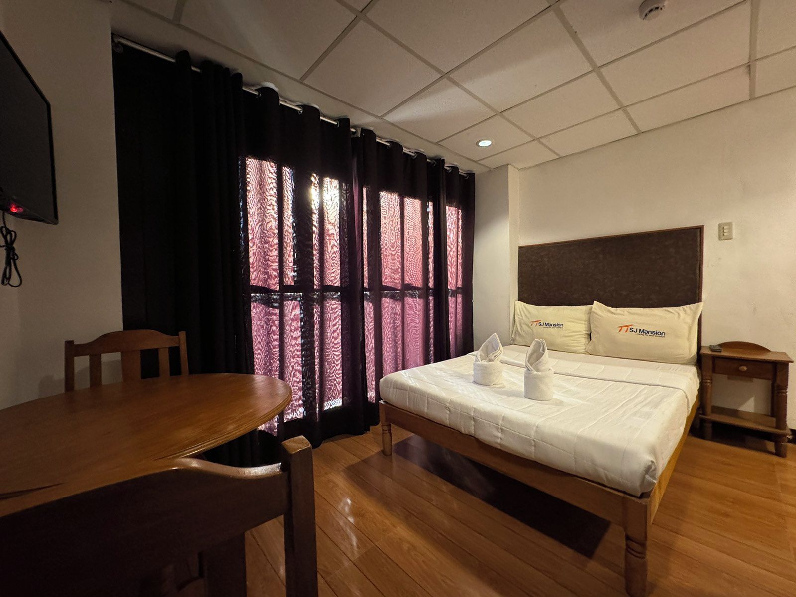 A hotel room with a wooden bed, a wooden table, and a window with patterned curtains.
