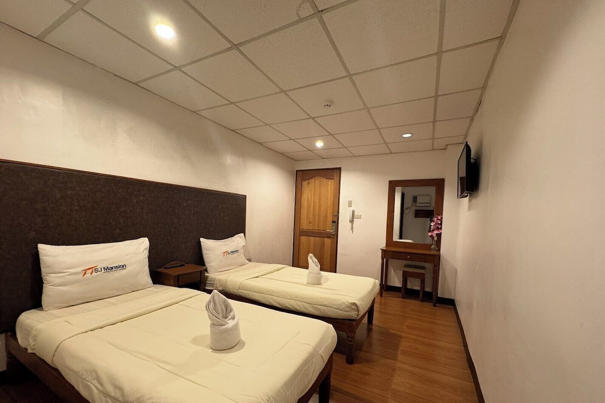 A hotel room with two beds, a wooden headboard, a small desk, a mirror, and a TV on the wall.