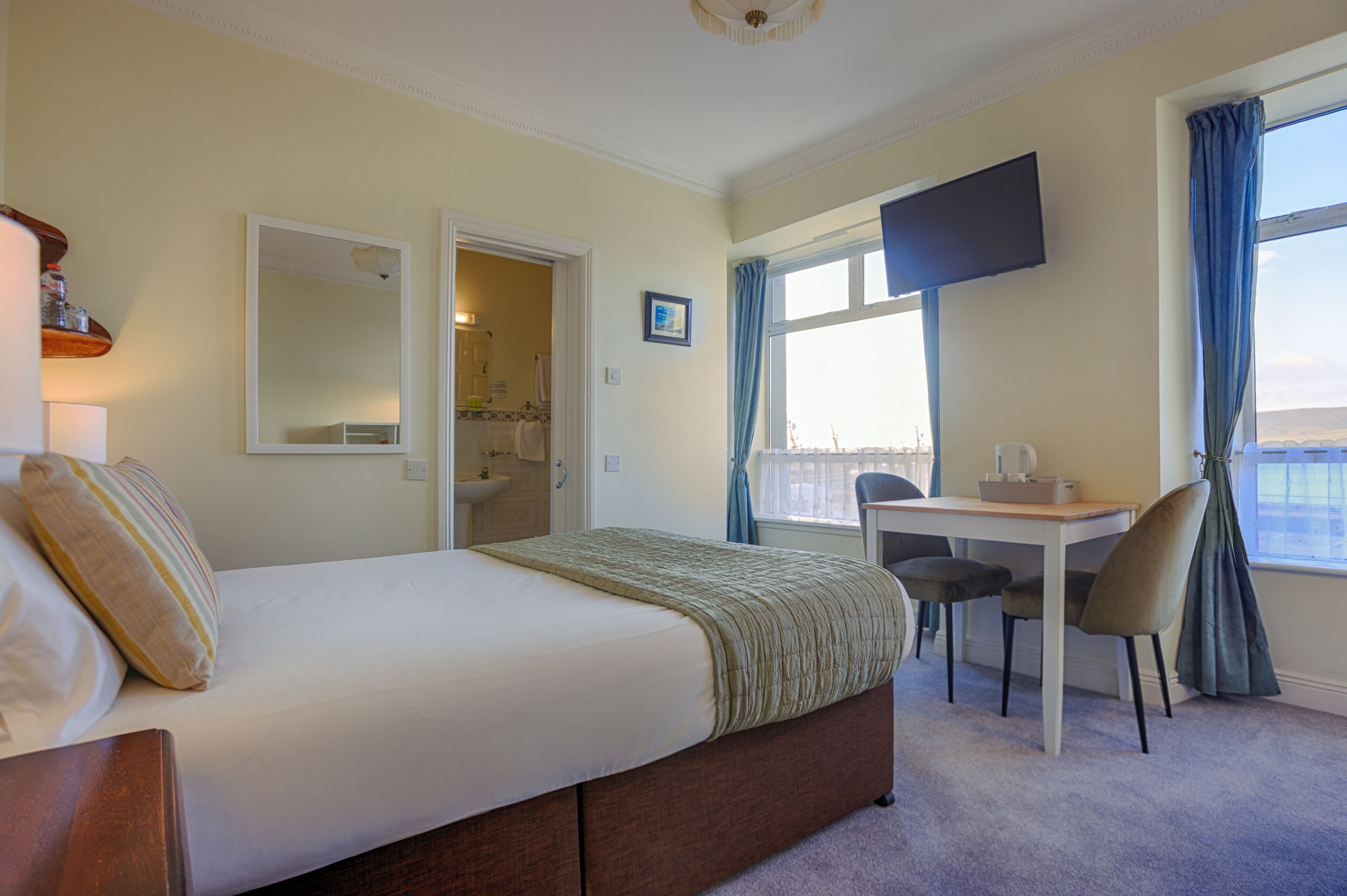 A bedroom with a bed, a desk, a chair, a TV, and a window with curtains.
