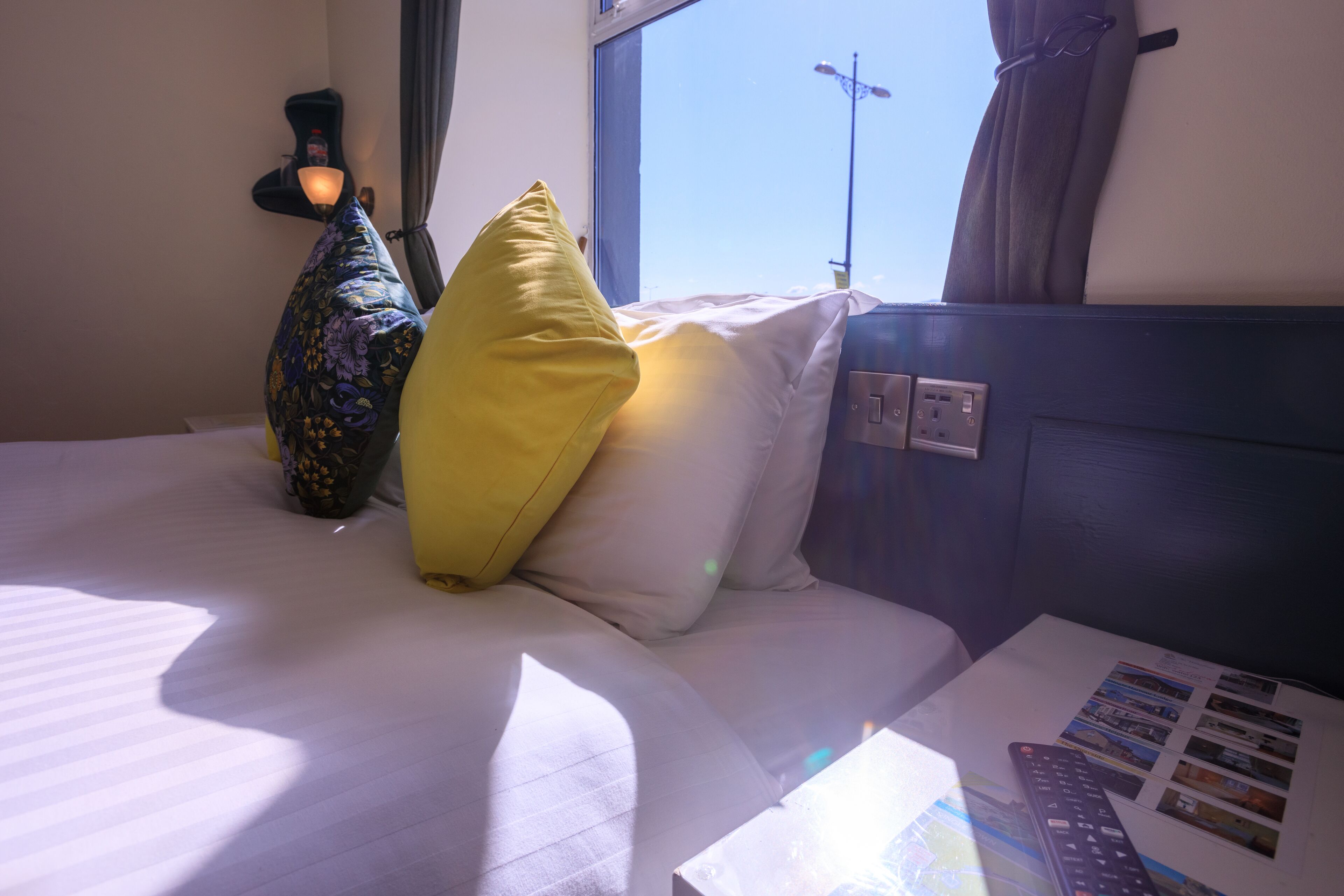 A hotel room with a bed, pillows, a lamp, a window, and a remote control on a table.