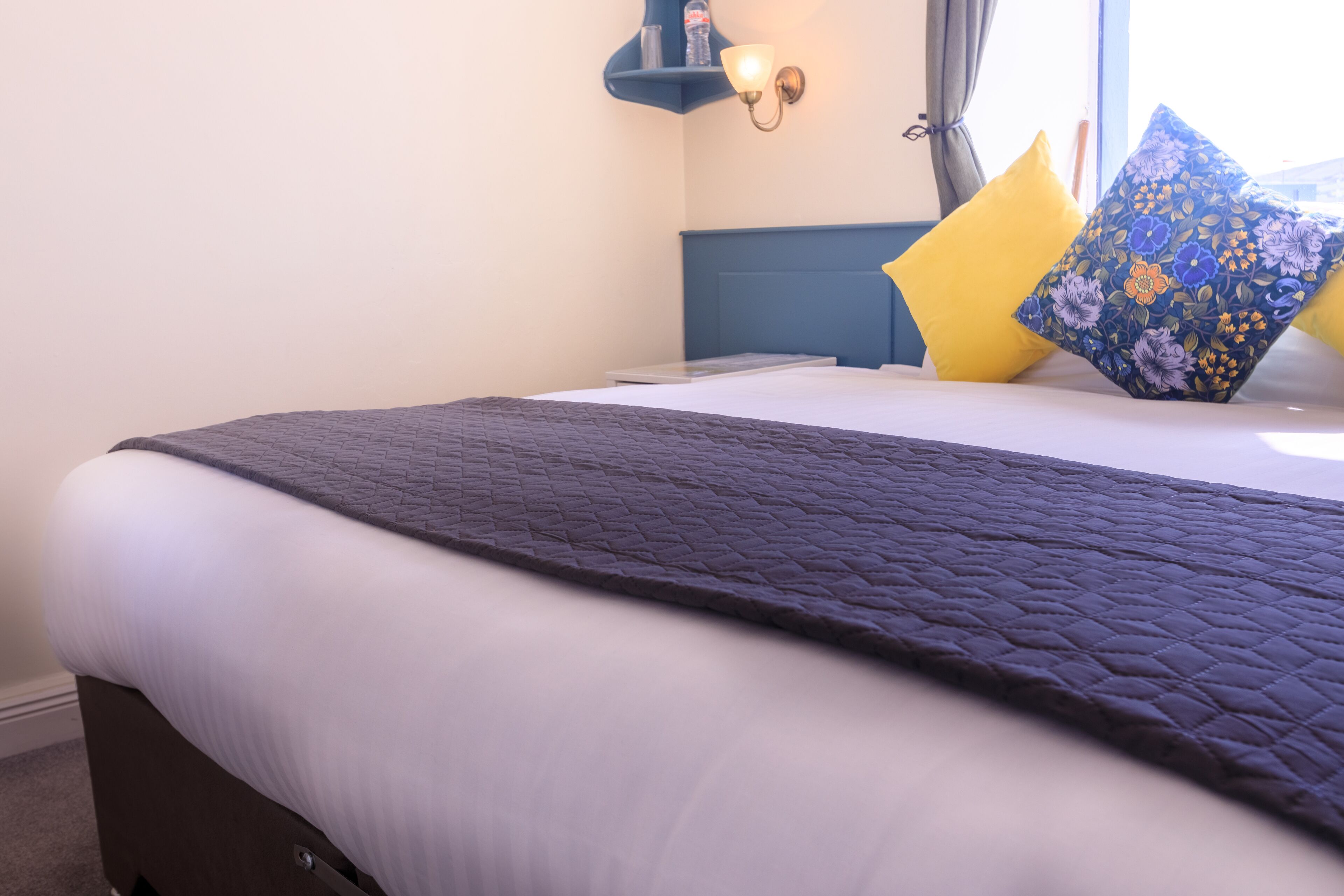 A neatly made bed with a quilted headboard, a bedside table with a lamp, and a window with curtains.