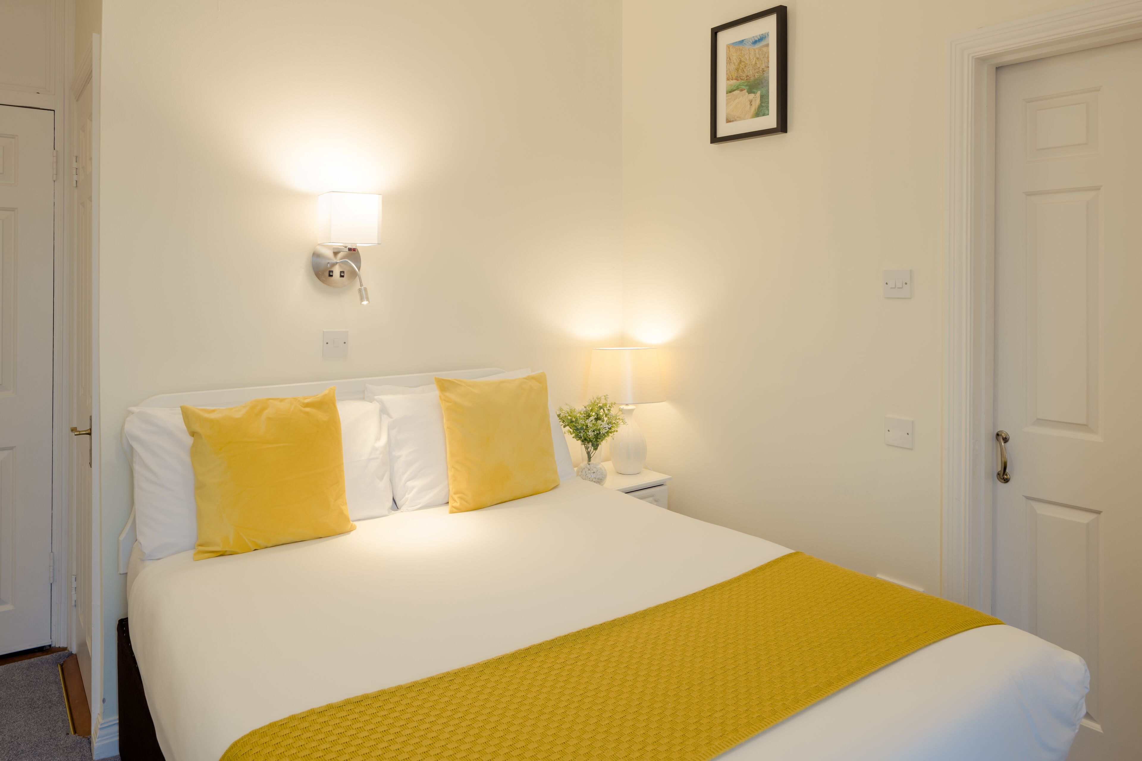A neatly made bed with white and yellow bedding, a bedside lamp, and a wall-mounted light fixture.