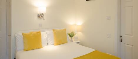 Una cama bien hecha con ropa de cama blanca y amarilla, una lámpara de noche y un aplique de pared.