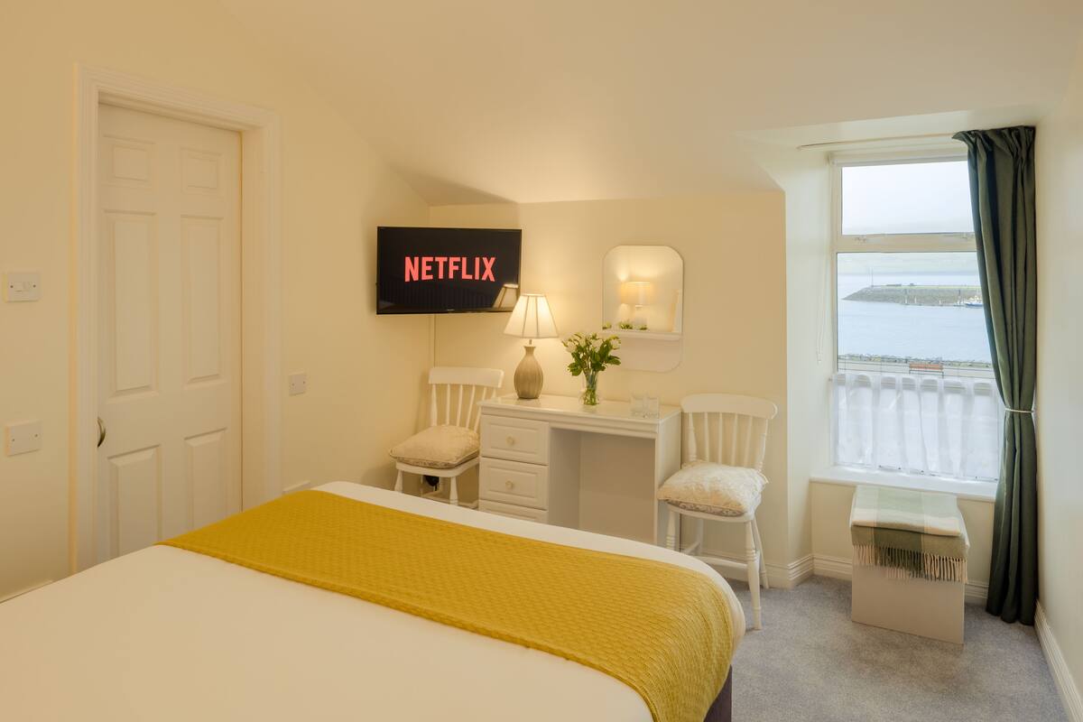 Habitación de hotel con cama, escritorio con silla, televisor montado en la pared, ventana con vista al mar y balcón con cortina.