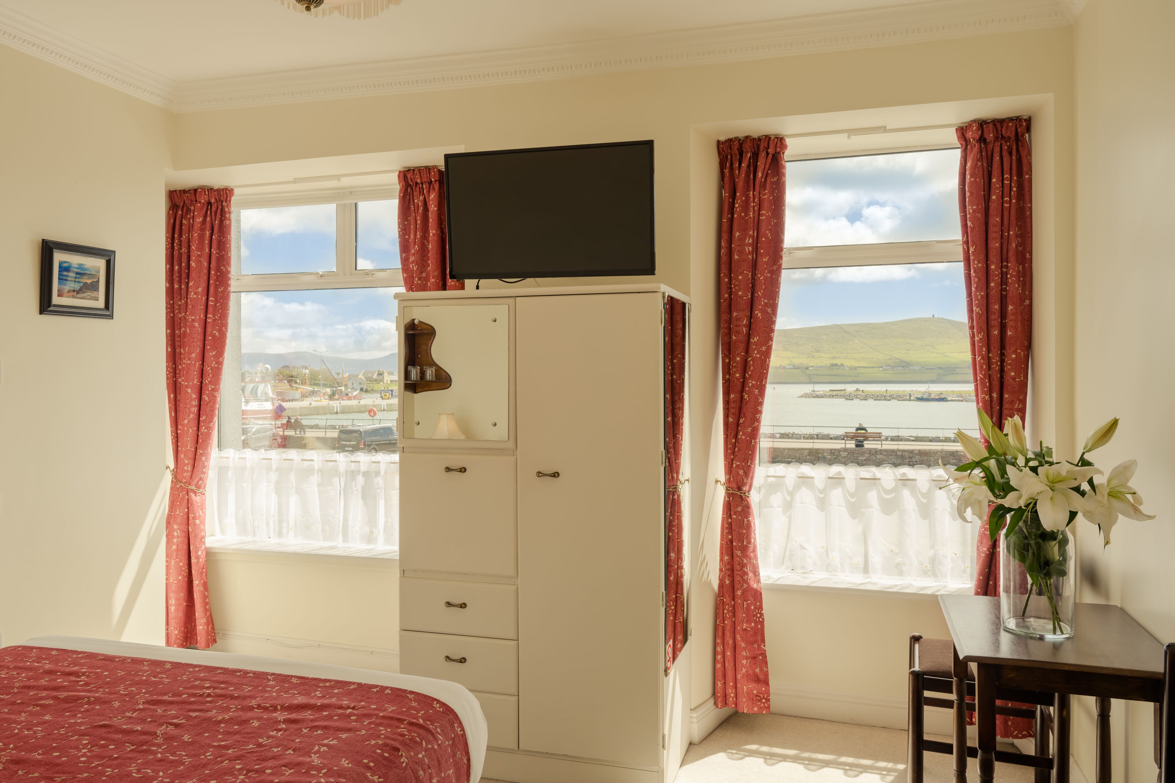 A room with a bed, a TV on a cabinet, a desk with a vase of flowers, and a window with curtains.