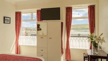 A room with a bed, a TV on a cabinet, a desk with a vase of flowers, and a window with curtains.