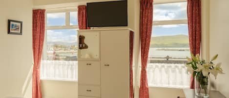 A room with a bed, a TV on a cabinet, a desk with a vase of flowers, and a window with curtains.