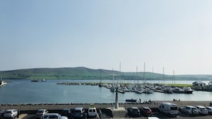 A marina with boats docked, a parking area with cars, and a grassy hill in the background. - The Waterfront (Dingle)
