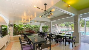 A covered outdoor dining area with multiple tables and chairs, a pool in the background, and a stone wall. - Sawasdee Place Hotel (Pattaya)