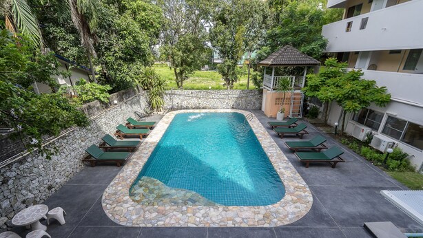A rectangular pool with a stone border, surrounded by lounge chairs and tables, set against a backdrop of greenery and a stone wall. - Sawasdee Place Hotel (Pattaya)