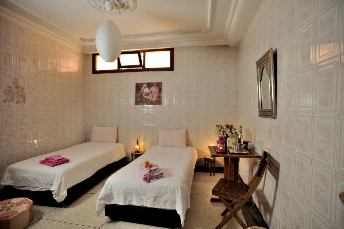 Une chambre d’hôtel avec deux lits, une petite table ornée d’un vase et de bougies, et une chaise en bois.