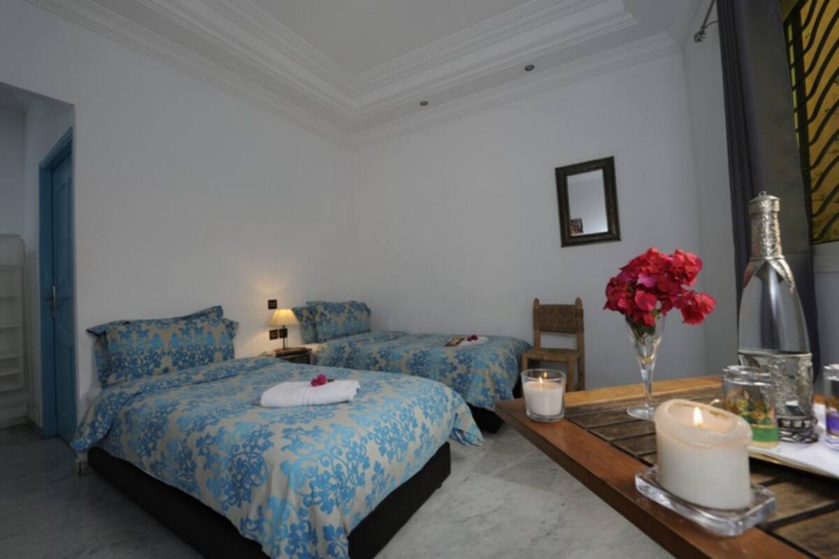 Une chambre d’hôtel avec deux lits, une table en bois sur laquelle est posée une bougie, un vase de fleurs et un miroir au mur.