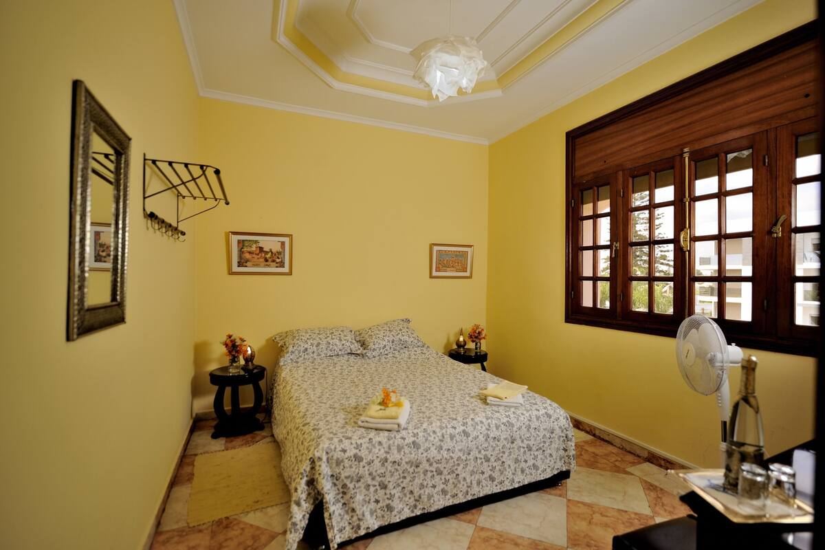 Une chambre à coucher comprenant un lit, une table de chevet avec une lampe, un ventilateur fixé au mur et une fenêtre avec des stores.