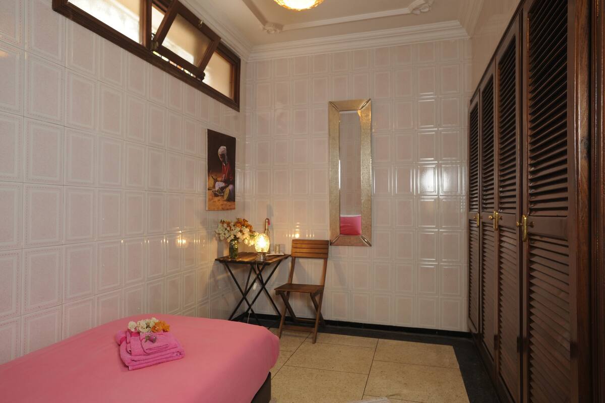 Une chambre avec un lit, une chaise, une petite table ornée de fleurs, un miroir et une armoire.