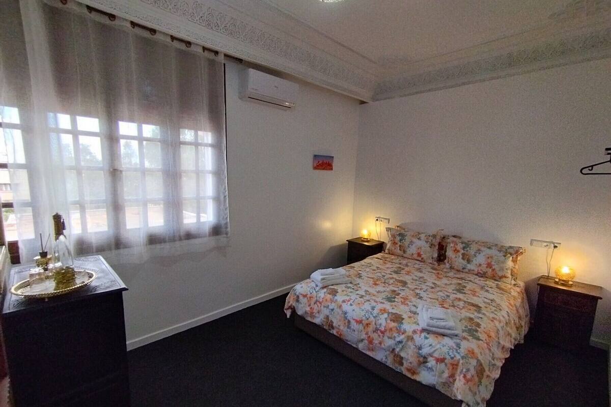Une chambre d’hôtel avec un lit, deux tables de chevet, une fenêtre avec des rideaux, un climatiseur et un petit drapeau accroché au mur.