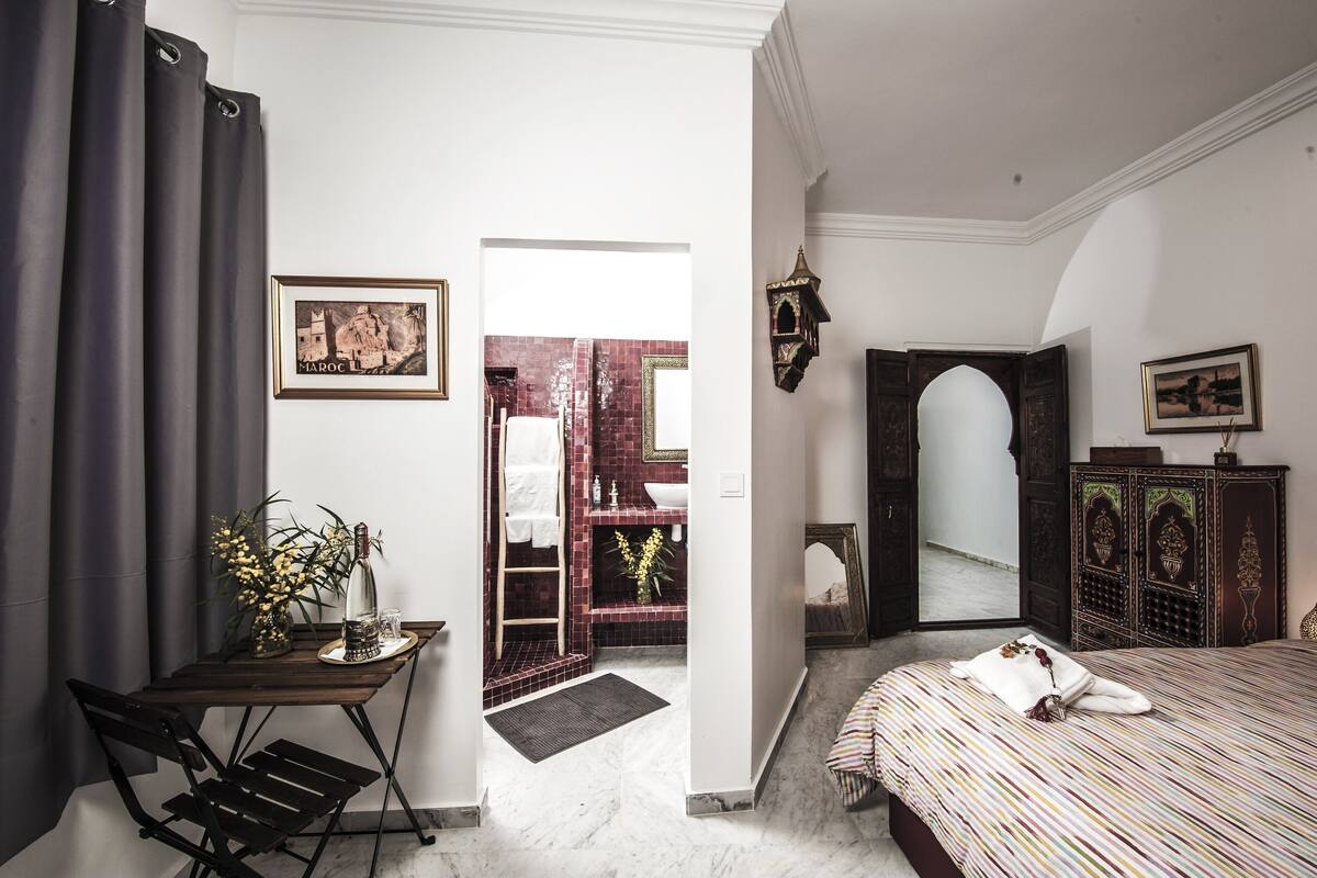 Une chambre d’hôtel avec un lit, un bureau agrémenté d’un vase et d’une bouteille, un miroir et une vue sur une salle de bain aux carreaux rouges.