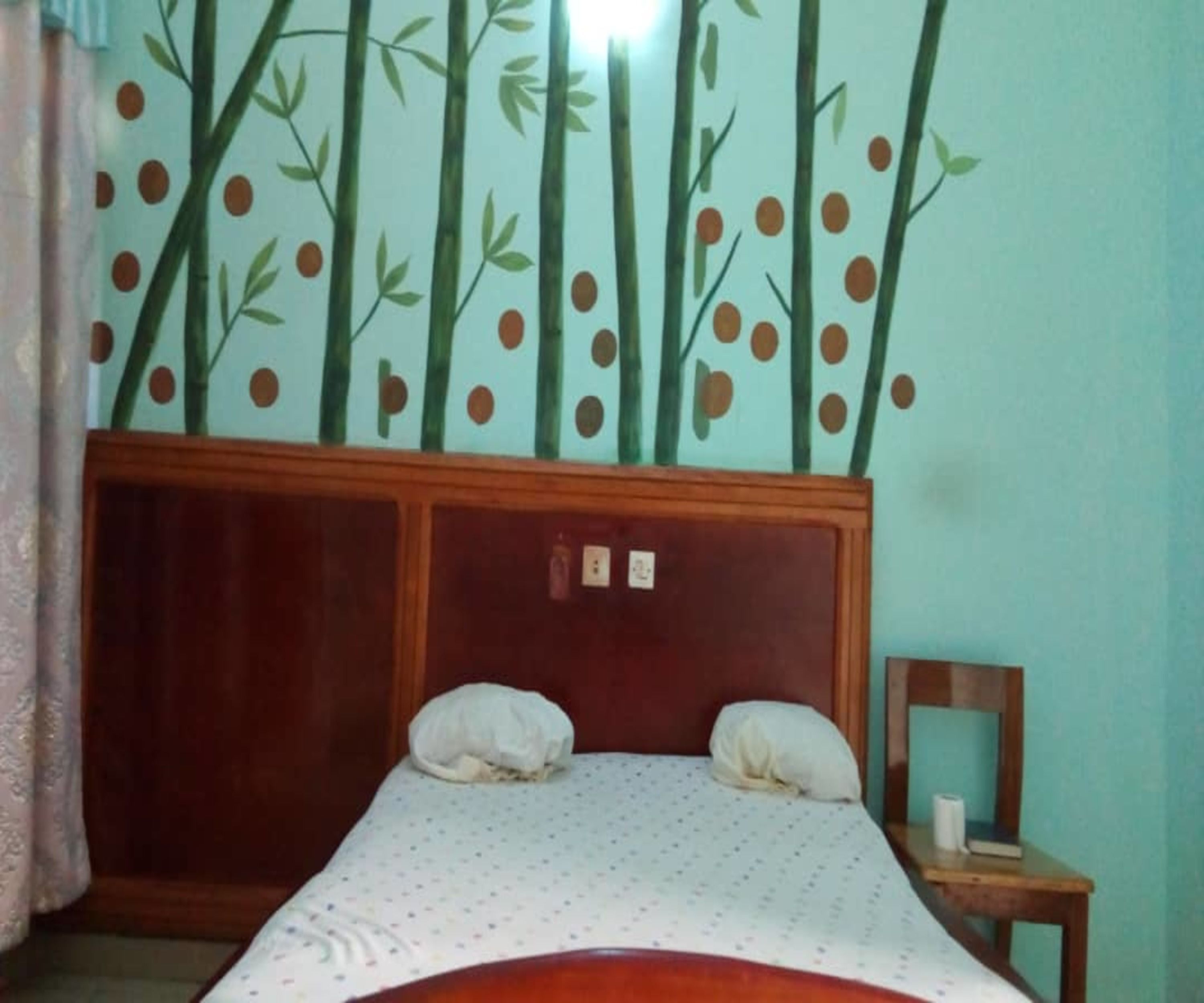 A bedroom with a wooden headboard, two pillows, a small bedside table with a lamp, and a wall with a bamboo and leaf pattern.