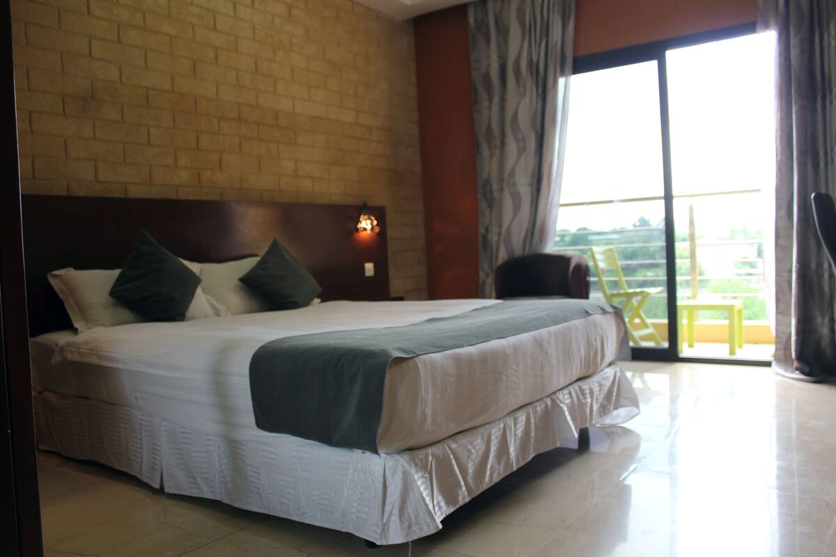 A hotel room with a large bed, a brick wall, a window with curtains, and a chair by the window.