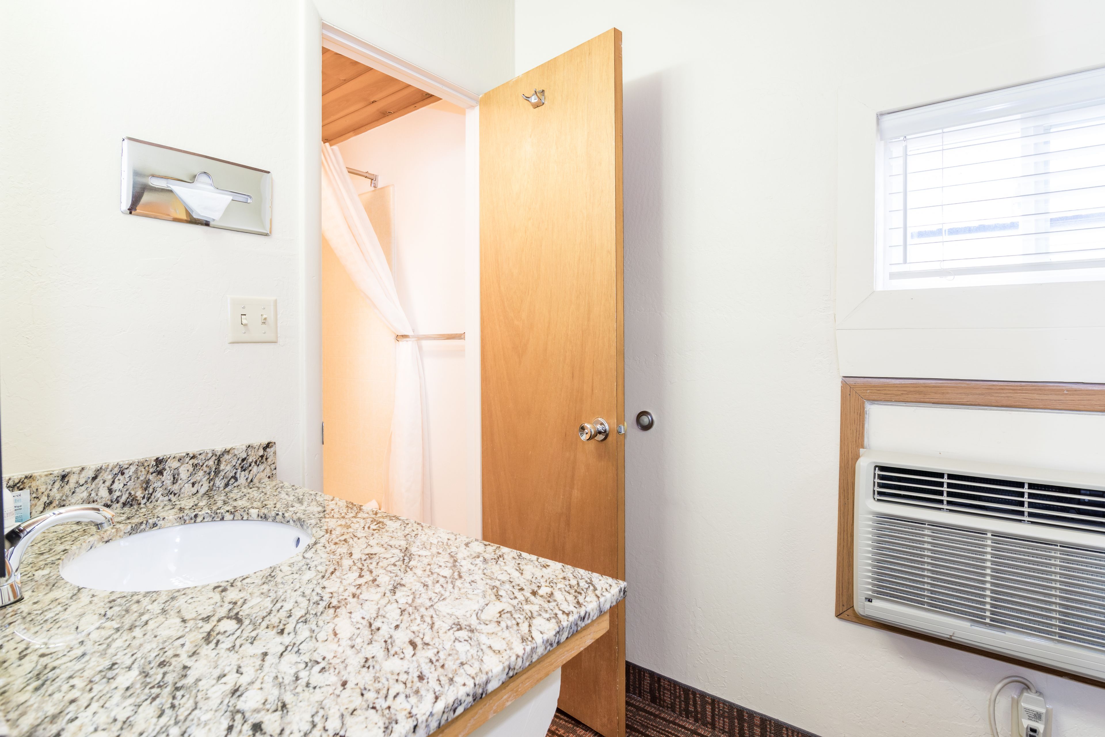 A bathroom with a granite countertop sink, a wooden door, a shower curtain, and a window with blinds.