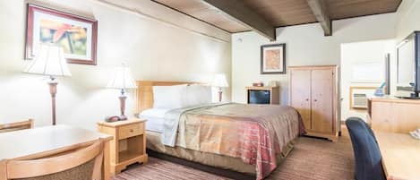 A hotel room with a wooden ceiling, a bed with a plaid blanket, a desk with a chair, a nightstand with lamps, and a wardrobe.