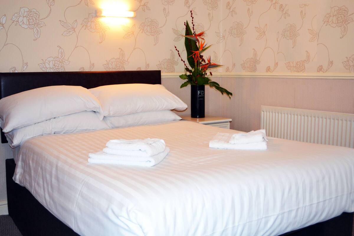 A neatly made bed with white linens, a dark headboard, and a vase with a floral arrangement on the nightstand.