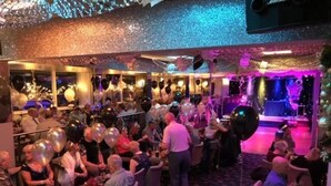 A crowded indoor event with people seated and standing, balloons, and a stage with performers. - Lyndene Hotel (Blackpool)