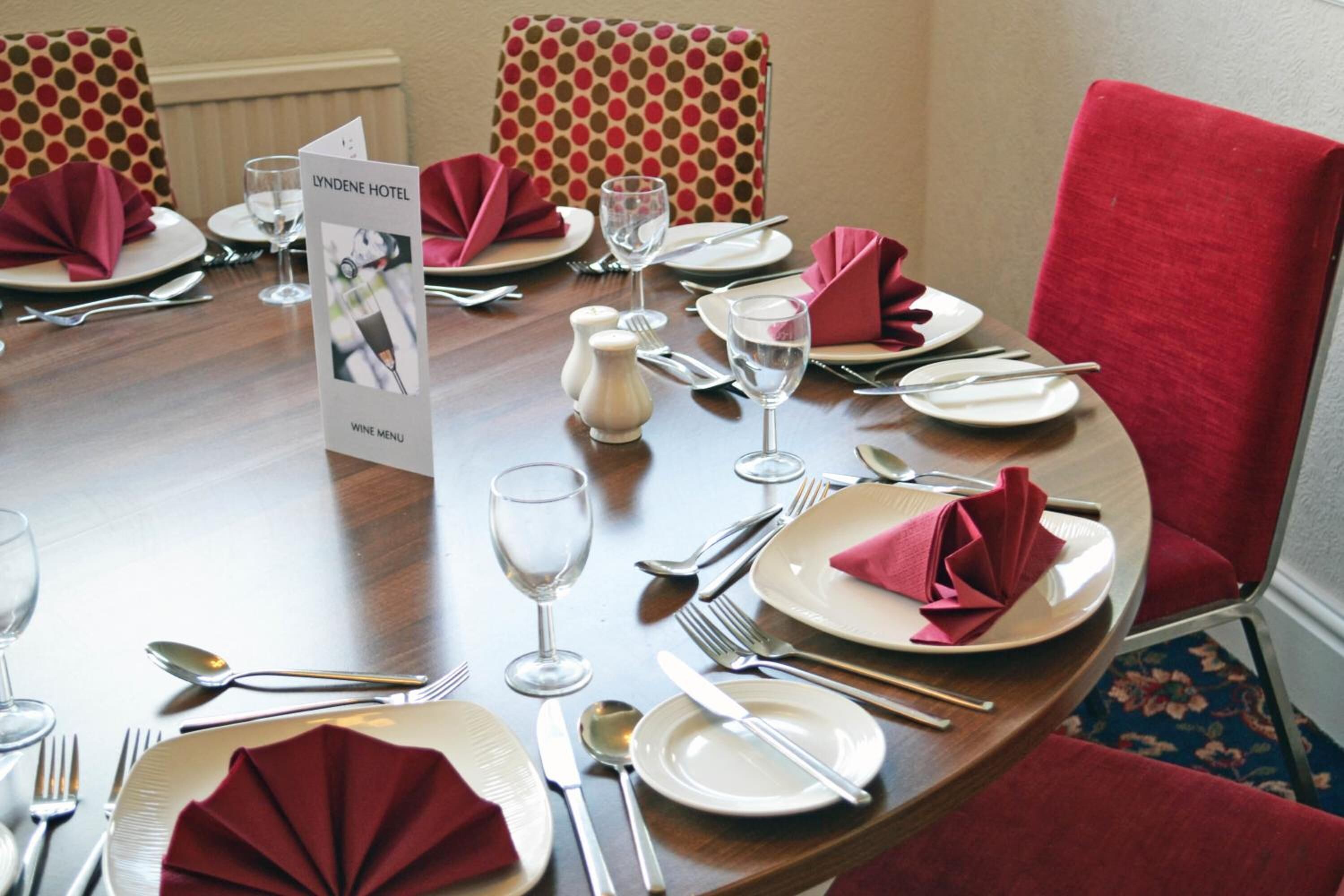 Een eettafel gedekt voor een maaltijd, met rode servetten, witte borden en bestek. Er ligt een wijnkaart op tafel.