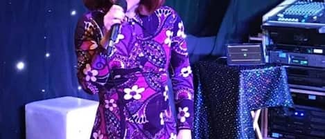 Una persona en el escenario con un micrófono, una mesa con equipo electrónico y un telón de fondo iluminado.